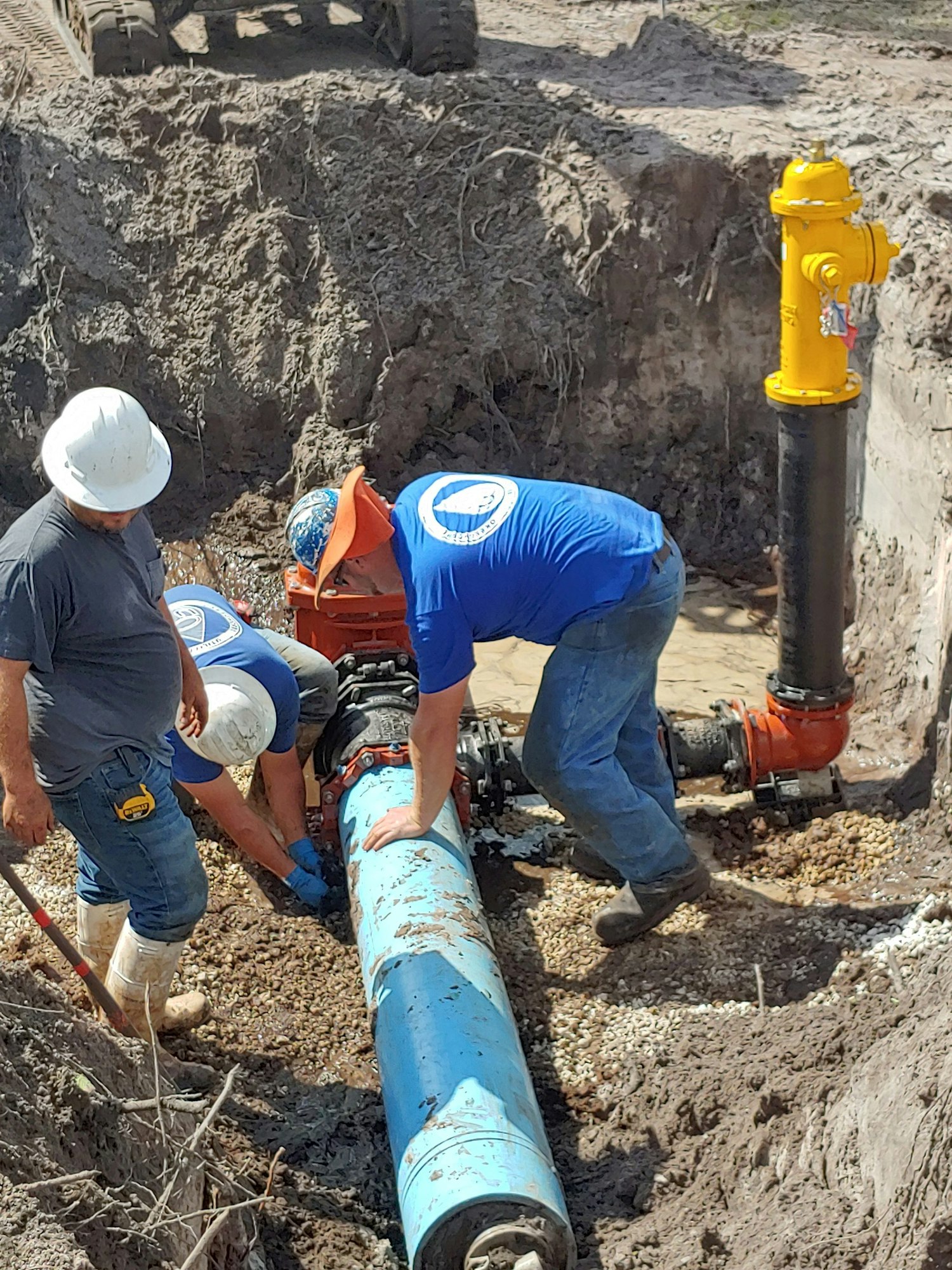 Workers installing a blue water pipe connected to a yellow fire hydrant in a muddy trench.