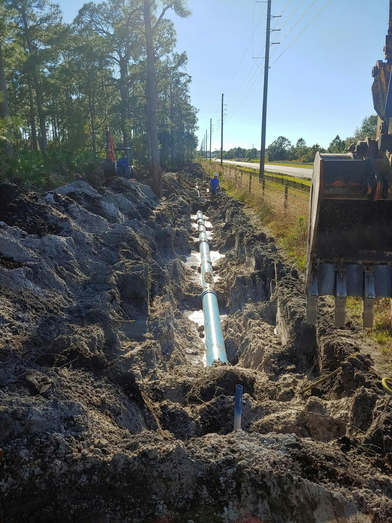 A trench with blue pipes being laid, surrounded by dirt and machinery, workers on site.