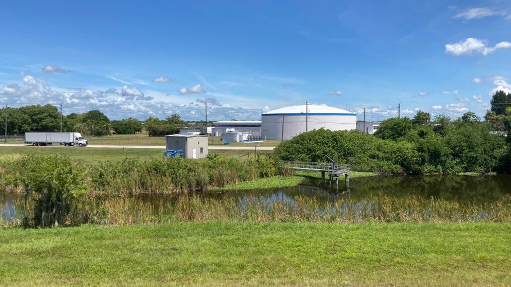 A field with greenery and a pond, a white tank structure, and a truck on a road under a blue sky.