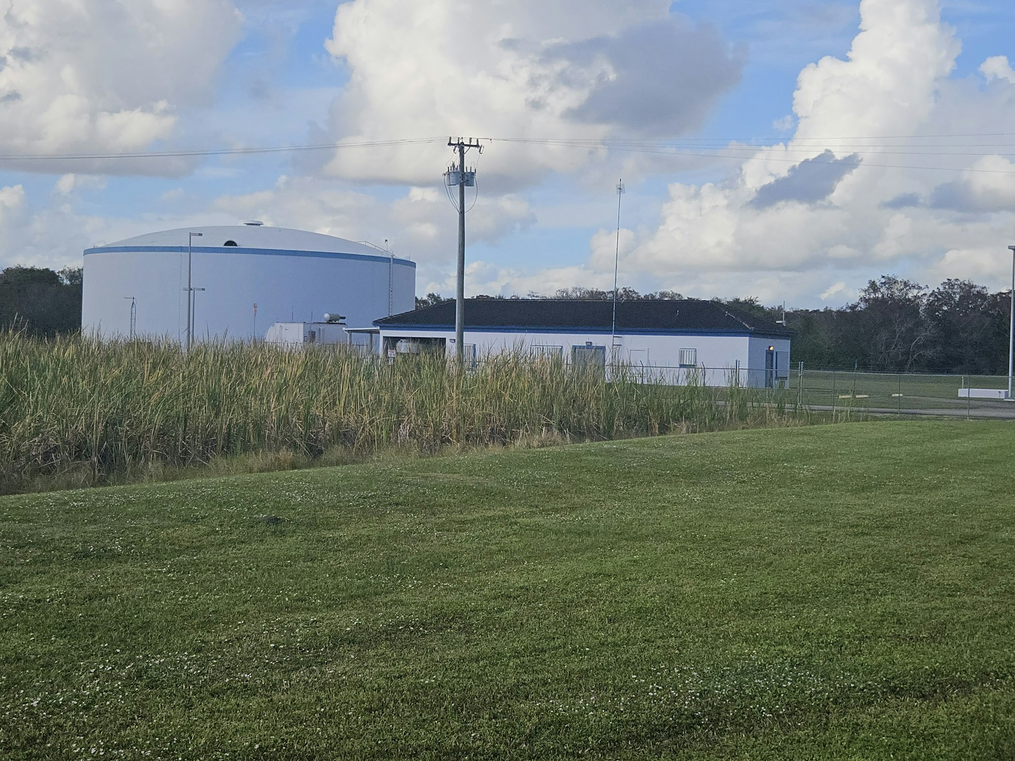 A large cylindrical tank and a small building in a grassy field under a partly cloudy sky.