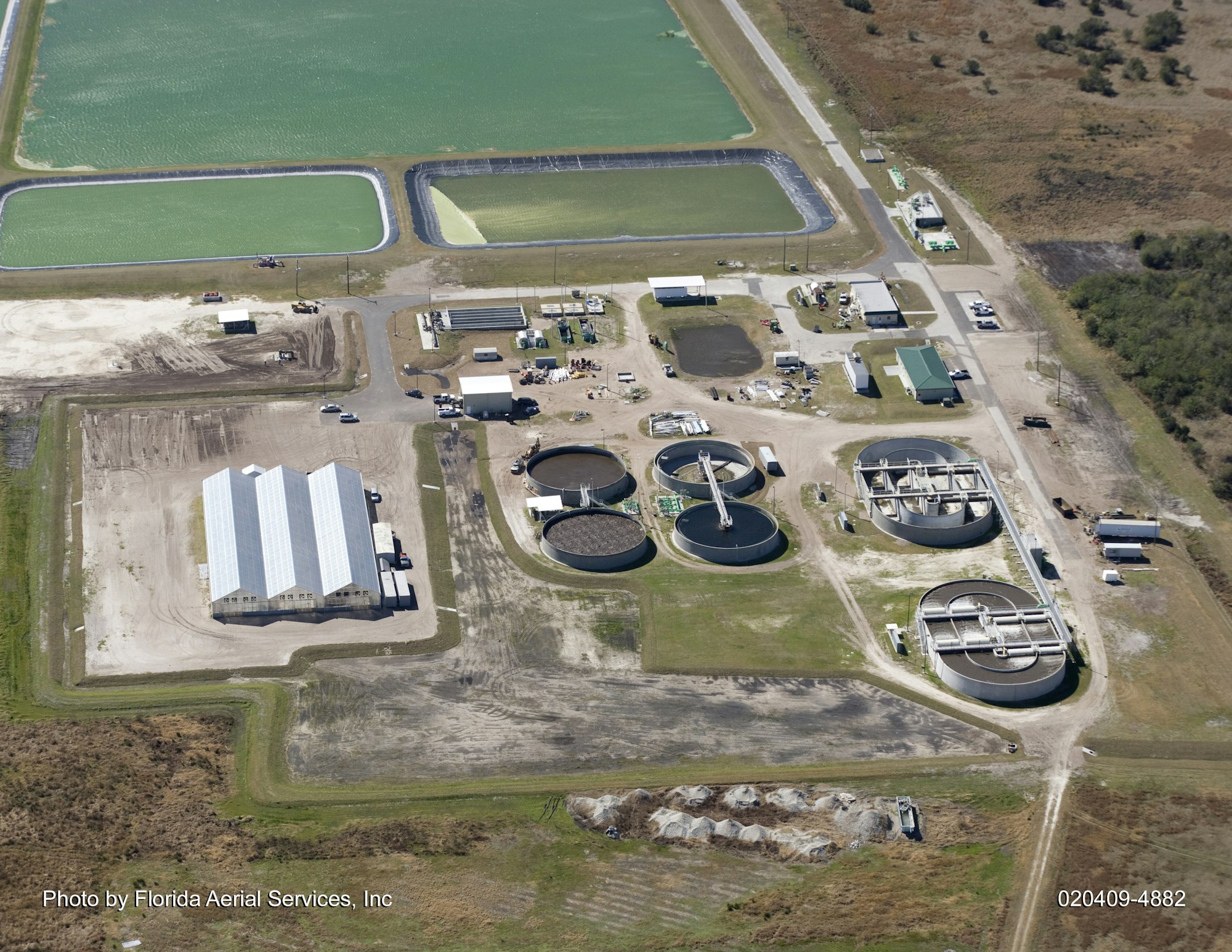 Aerial view of a wastewater treatment plant with multiple tanks, buildings, and nearby green pools.