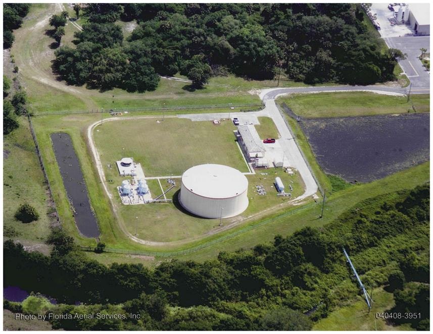 Aerial view of a large circular storage tank and adjacent facilities surrounded by greenery and access roads.