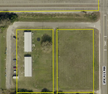Aerial view of two rectangular plots: one with buildings and vehicles, the other vacant, next to railway tracks and a road.
