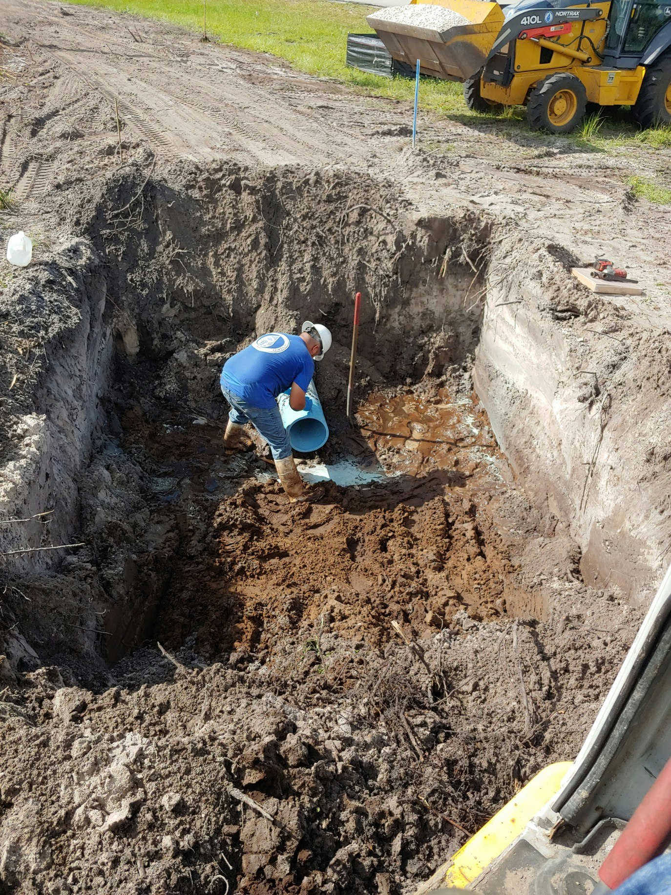 A worker is handling a pipe in a muddy trench. There's a backhoe nearby on the dirt road.
