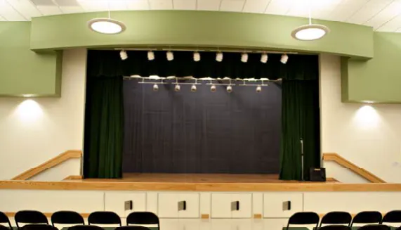A simple theater stage with green curtains and seating. Bright lighting fixtures overhead.