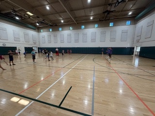 A gymnasium with Kids and Camp Counselors engaged in playing Dodge Ball on a wooden floor, with high ceilings and lined walls.