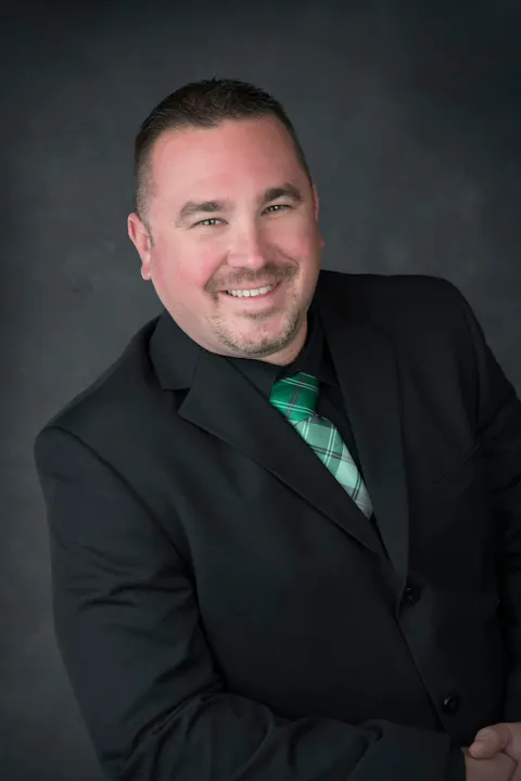 A man in a black suit with a green plaid tie smiles at the camera, set against a dark background.