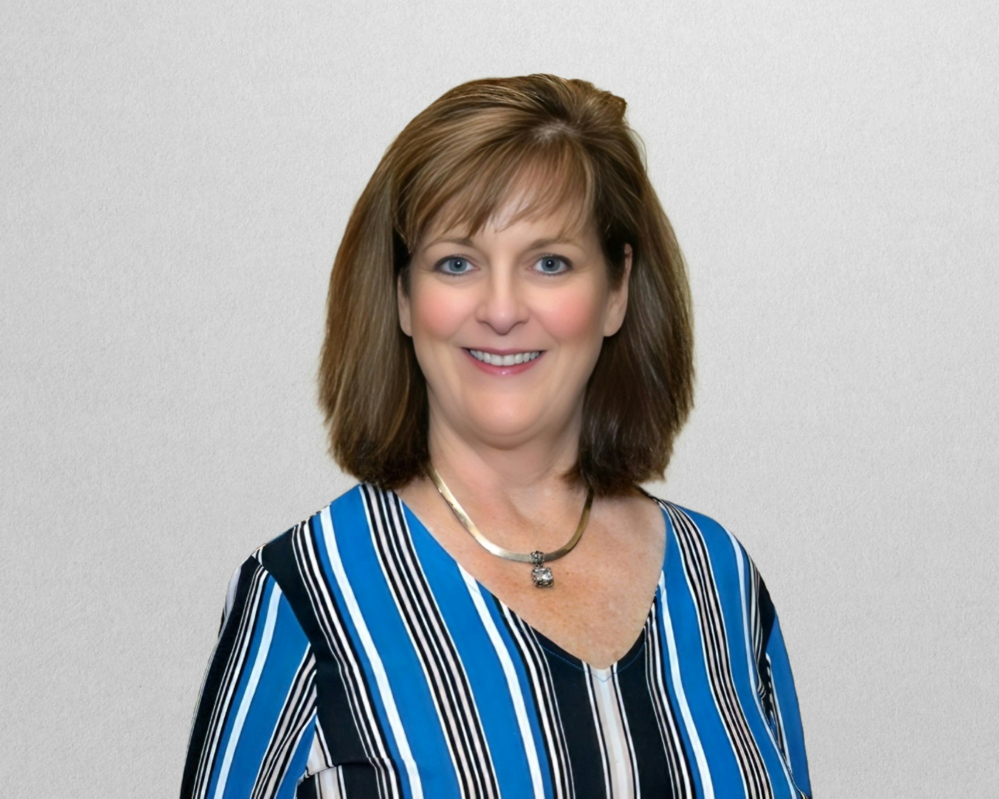 A woman smiling, wearing a blue and white striped shirt and a necklace, against a plain background.